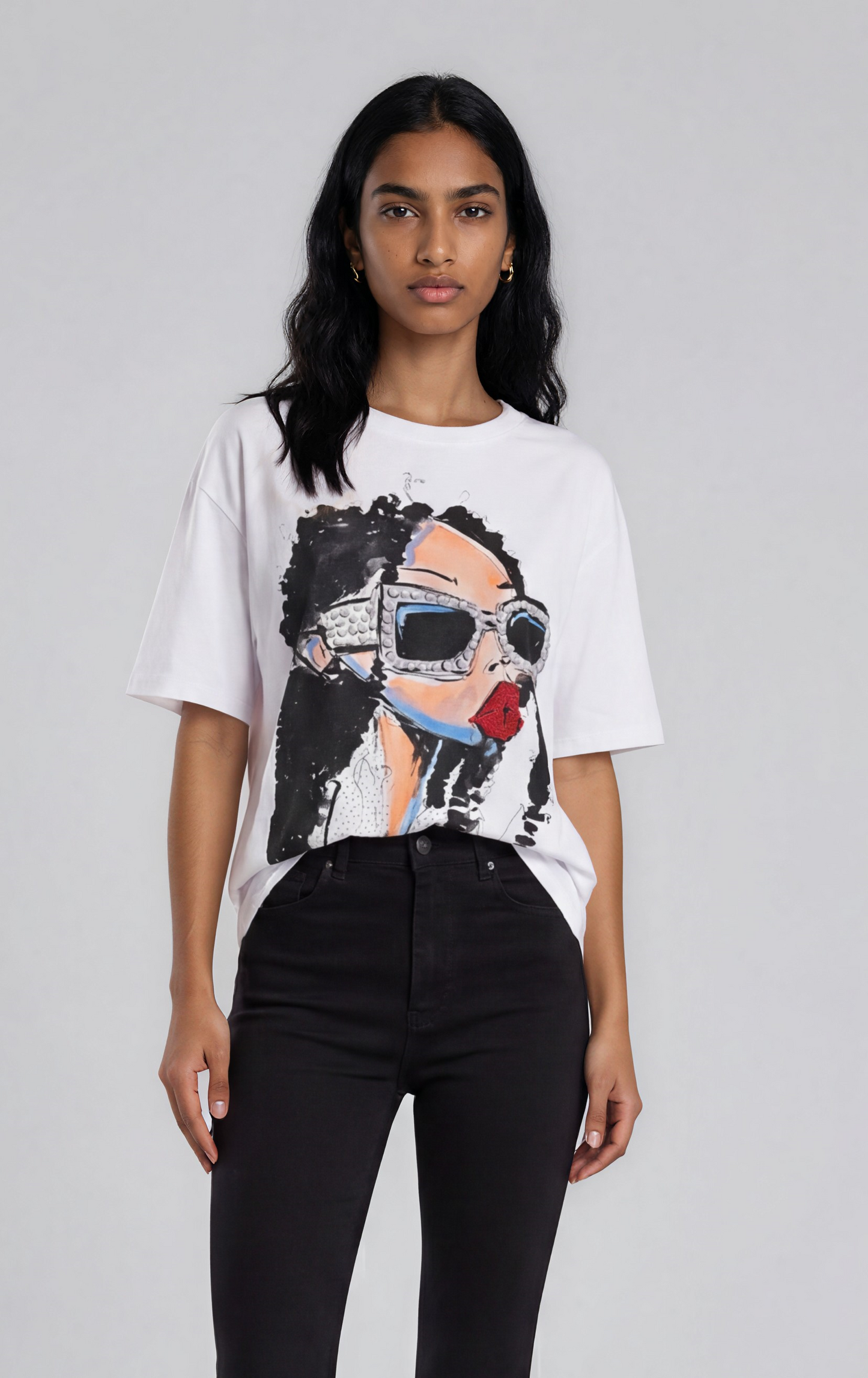 Woman wearing a white oversized graphic tee with a studded sunglasses face illustration paired with black pants in a studio setting – For Her NYC