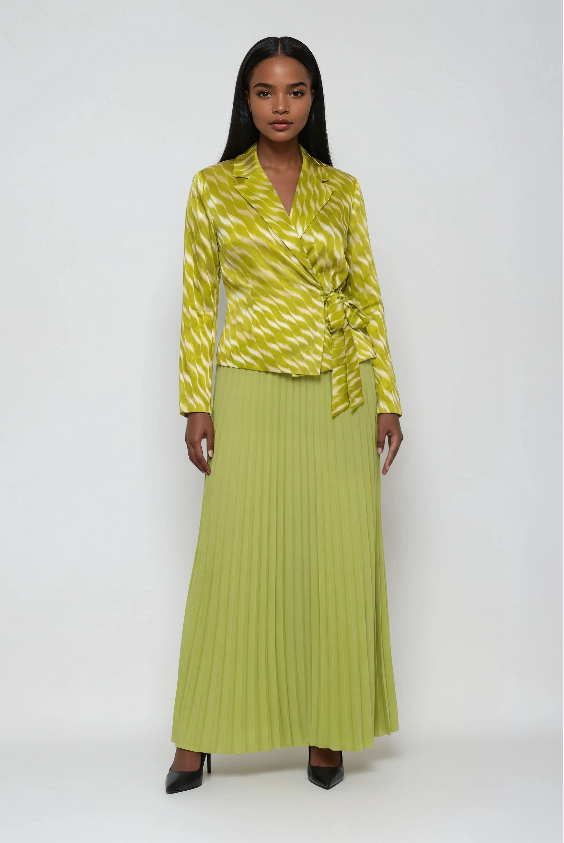 83009 yellow Woman wearing a yellow pleated midi skirt with a matching structured wrap-style jacket and tie waist, styled with heels — For Her NYC
