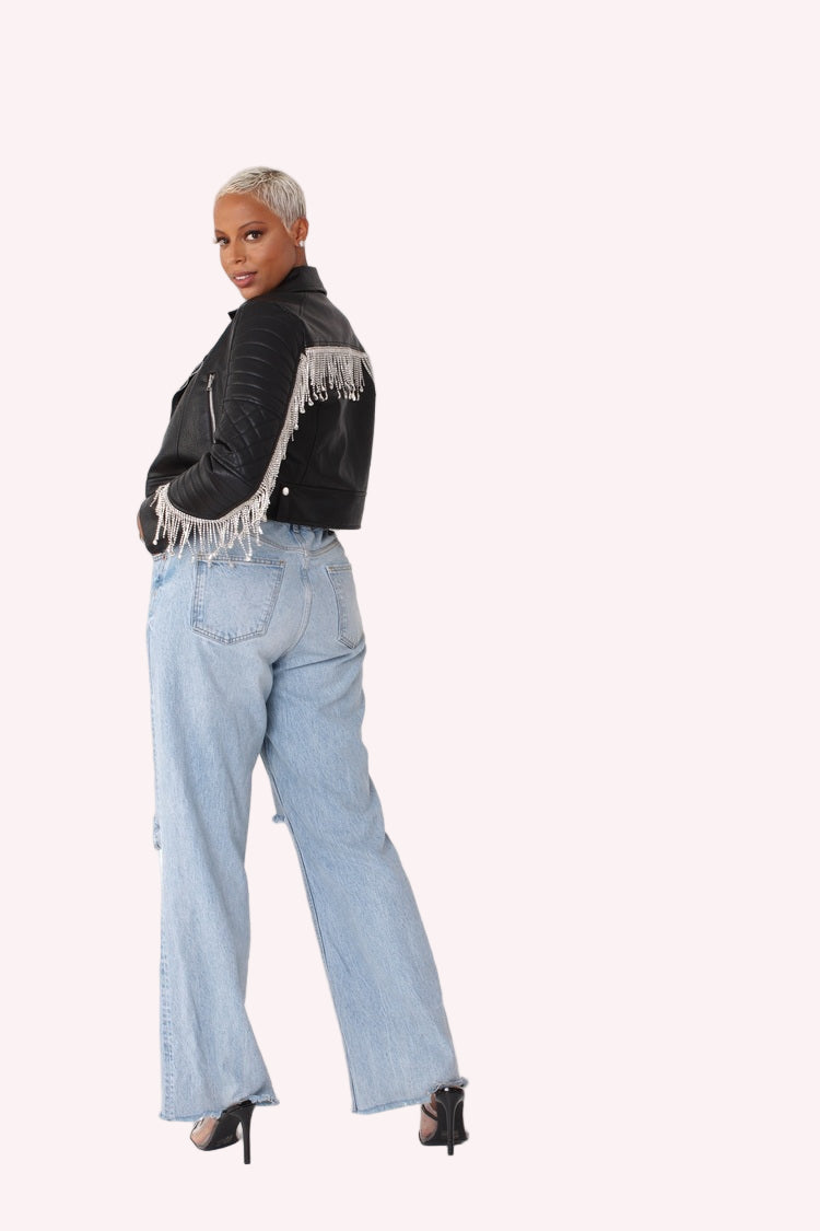 Back view of a black cropped faux leather jacket with fringe detailing, shown in an over-the-shoulder pose highlighting the draped fringe across the back, quilted sleeves, and cropped silhouette styled with light denim by For Her NYC