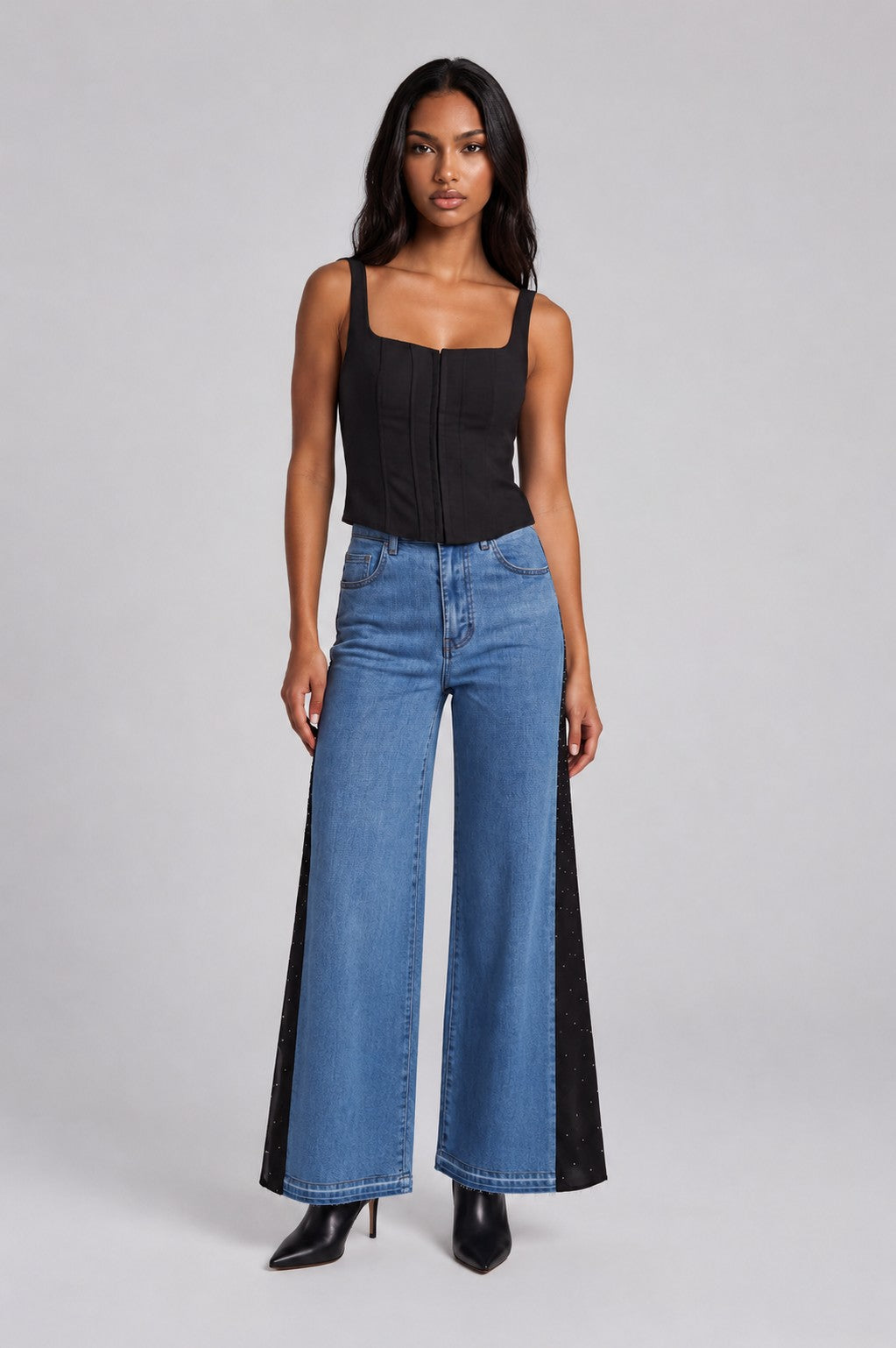 Model wearing the Crystal Drape Panel Denim Jeans featuring a medium wash denim front, black crystal-embellished contrast drape panels, a high-rise waist, and a wide-leg silhouette paired with a black camisole. For Her NYC.