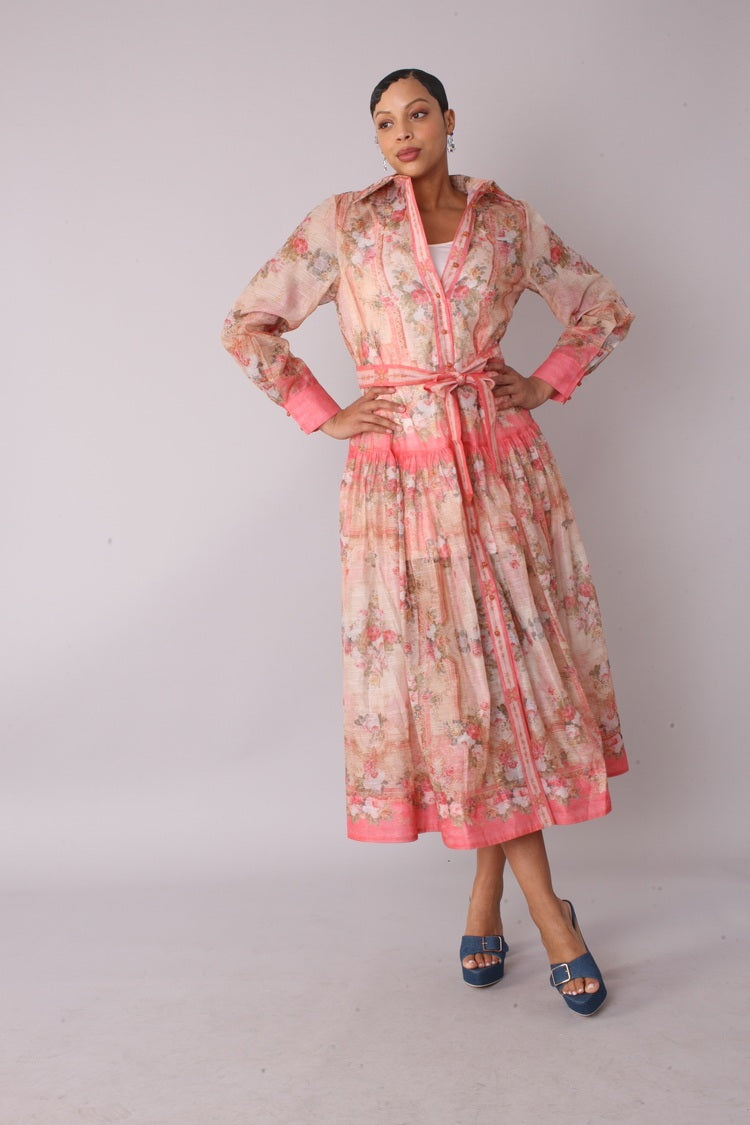 Front view of the pink floral print shirt dress with long sleeves, collared neckline and waist tie belt, model standing hands on hips wearing blue heels.