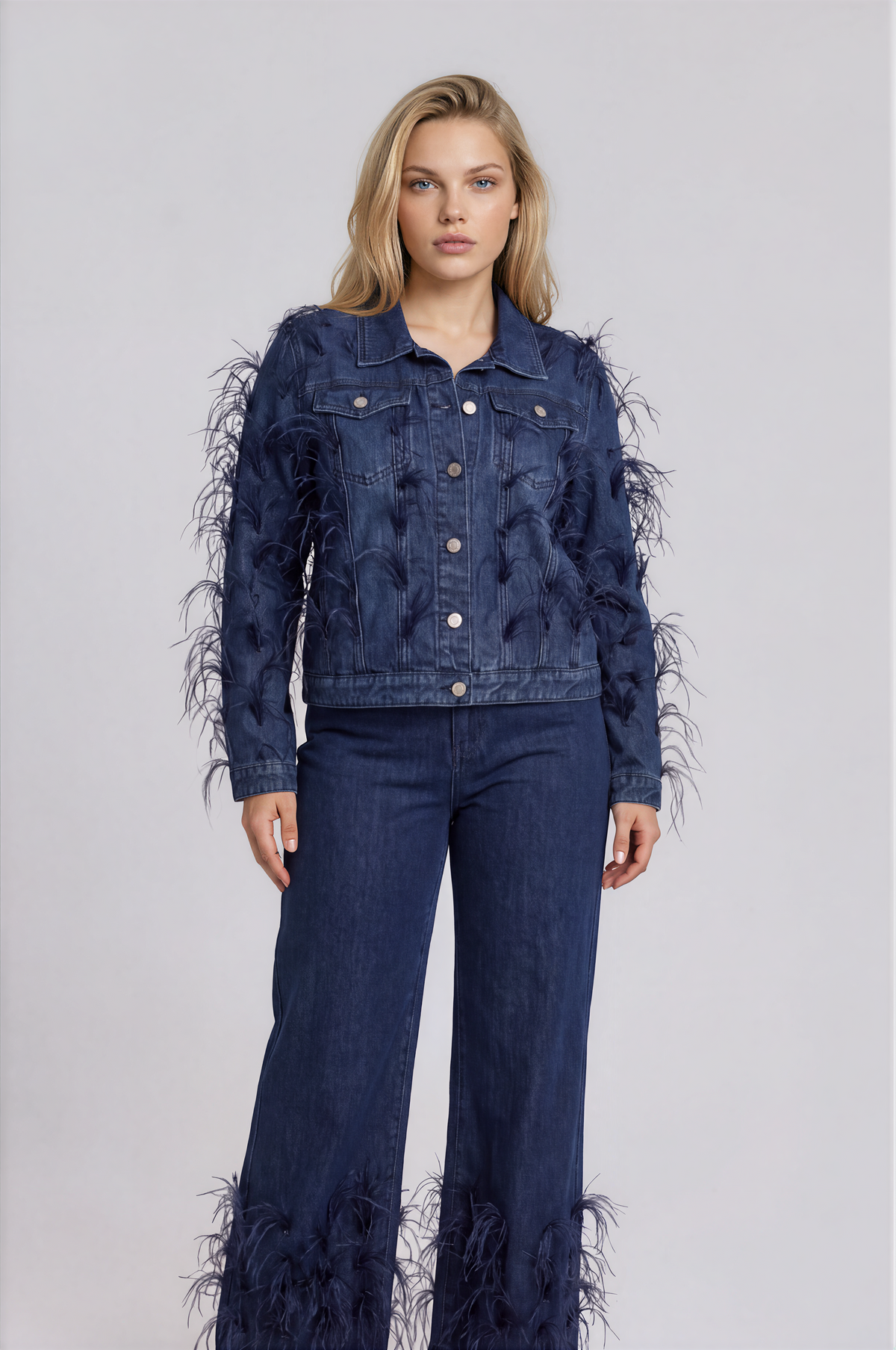 Three-quarter front view of a model wearing the dark-wash For Her NYC Feather Trim Denim Jacket - 82303 with feather embellishments across the front and sleeves
