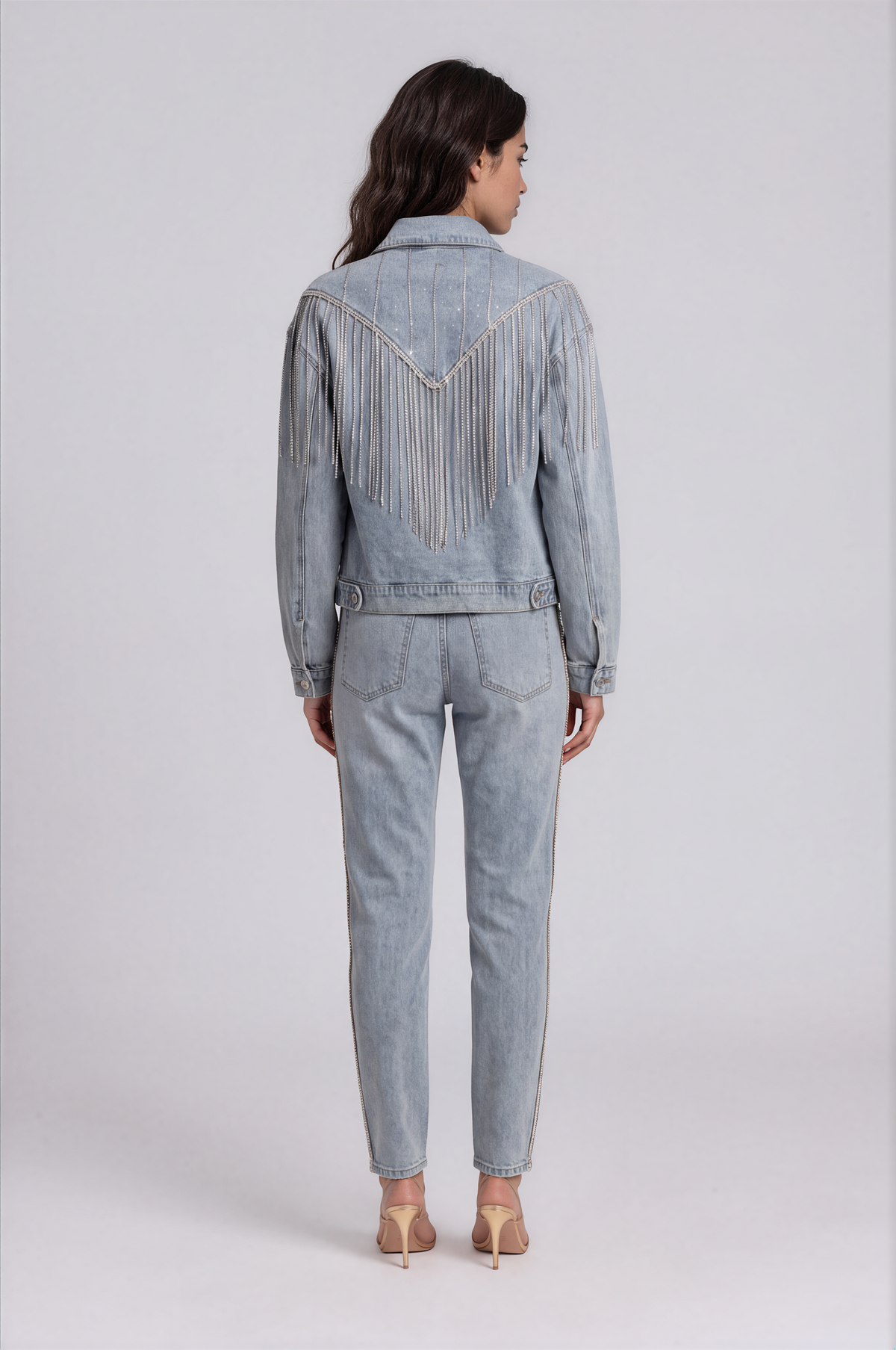 Back view of a model wearing the light-wash For Her NYC Rhinestone Fringe Denim Jacket - 82938 with a V-shaped yoke and cascading rhinestone fringe