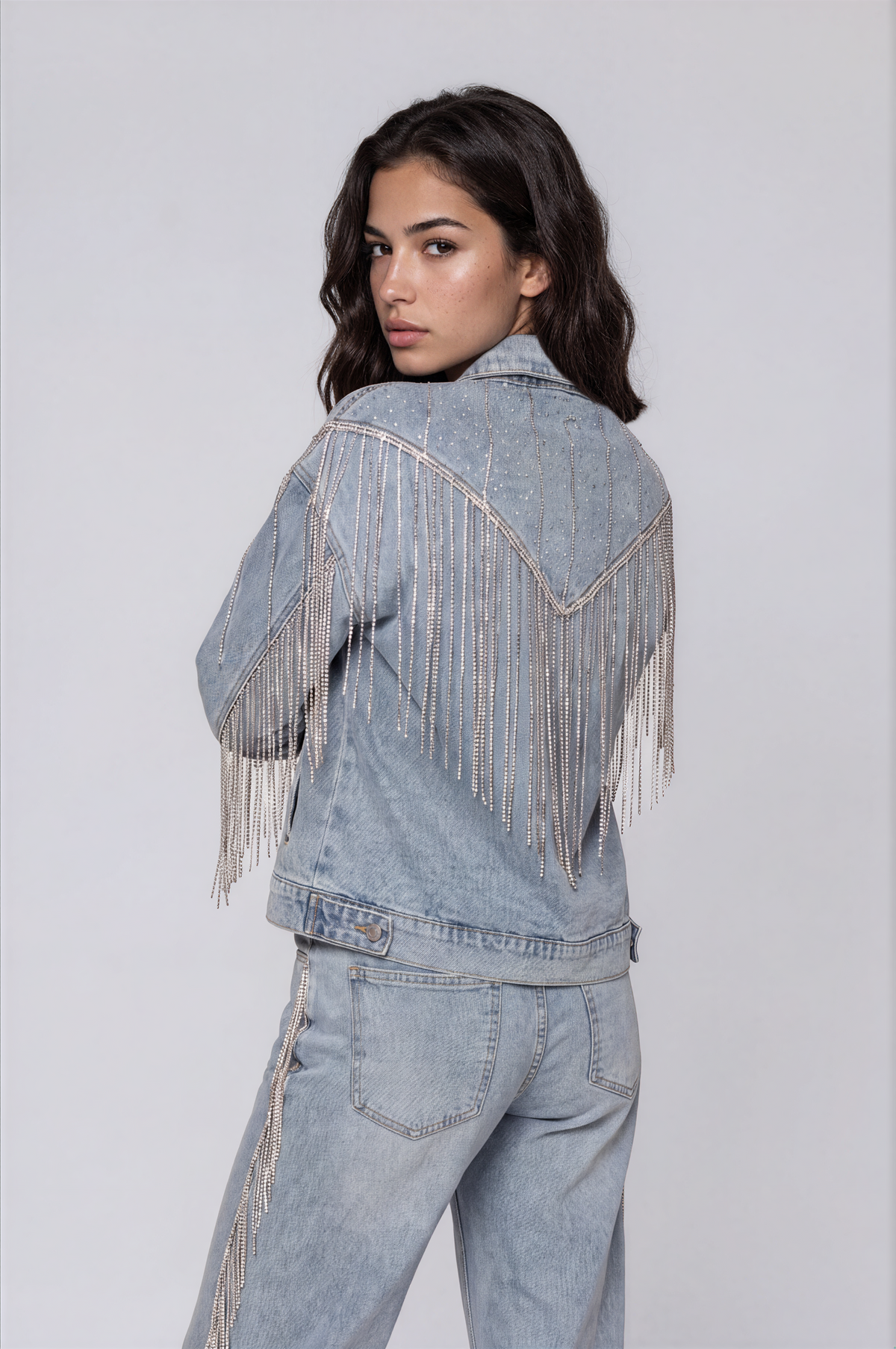 Three-quarter back view of a model wearing the light-wash For Her NYC Rhinestone Fringe Denim Jacket - 82938 with rhinestone embellishments and cascading fringe across the back and sleeves