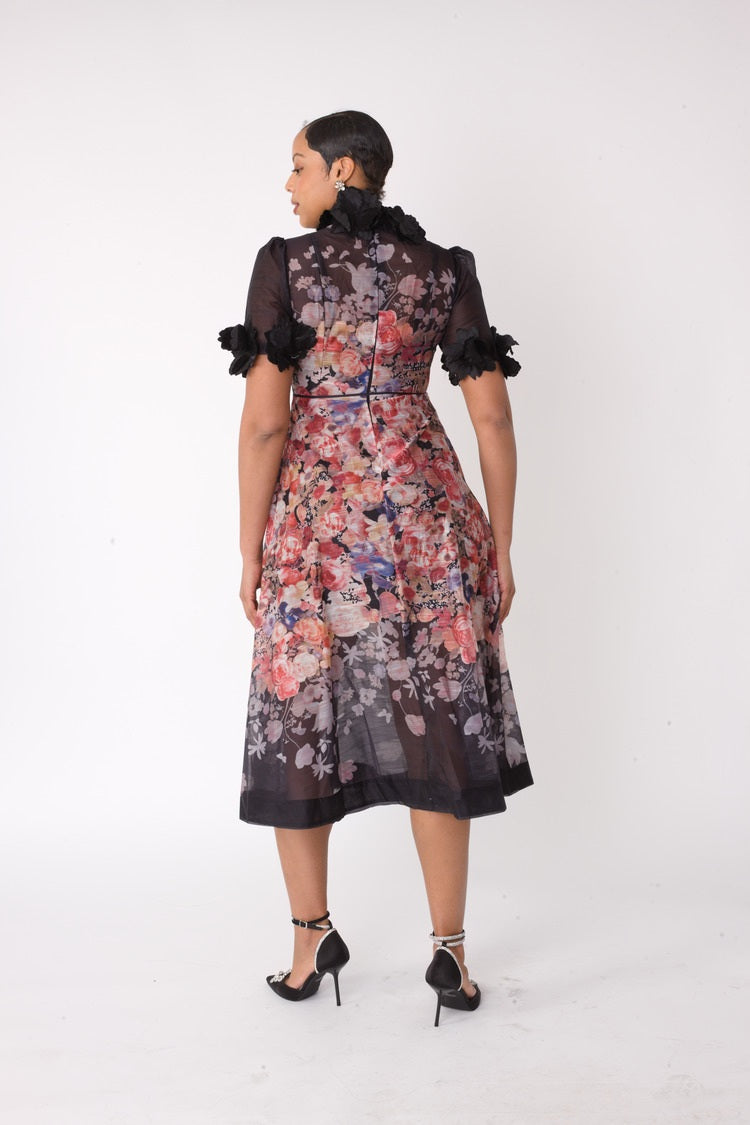 Back view of model wearing the black multi romantic floral midi dress, showcasing sheer organza upper bodice, floral print overlay, and elegant midi length silhouette by For Her NYC