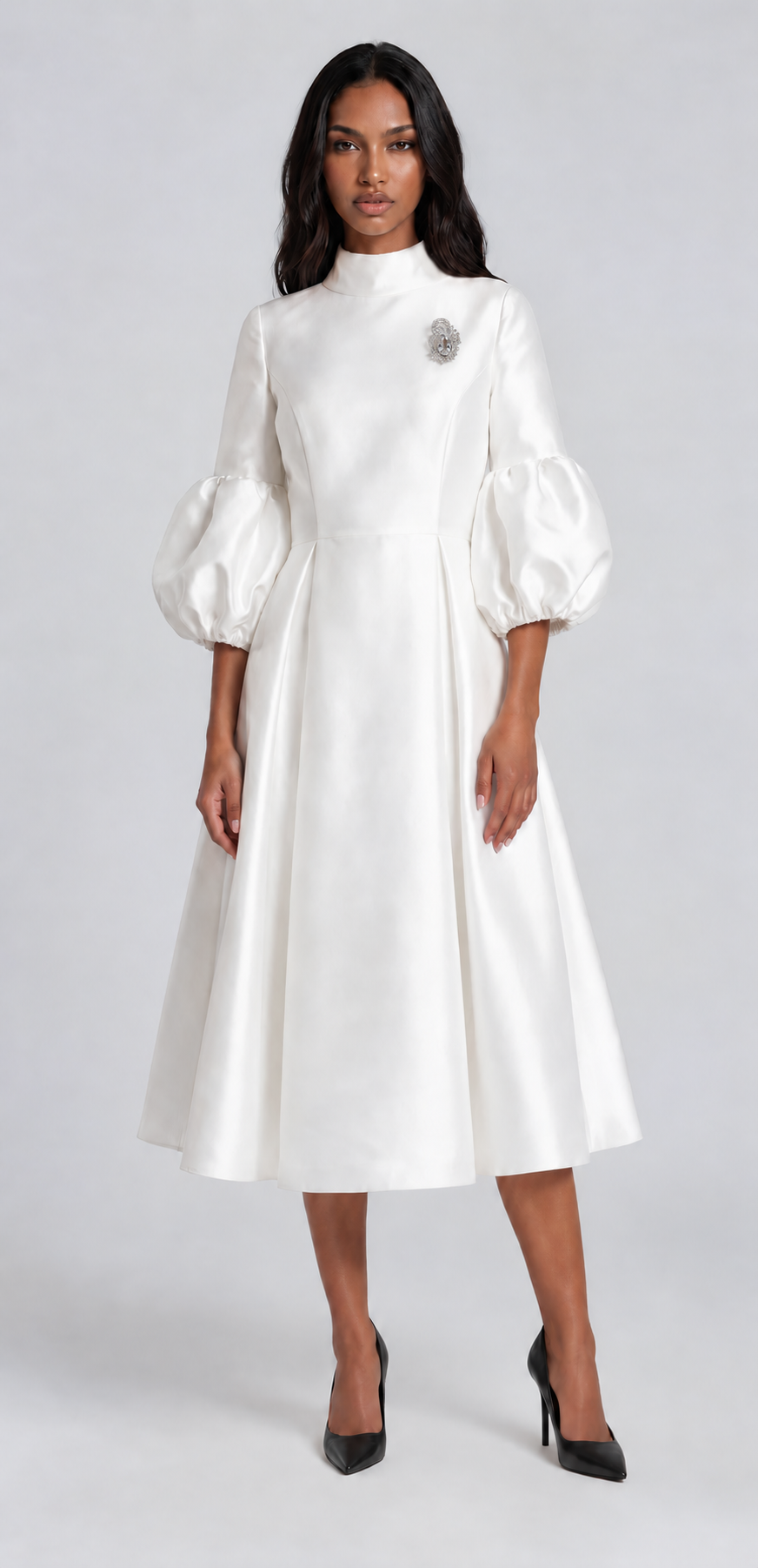 Woman wearing a white satin midi dress featuring dramatic puff sleeves, a modest high neckline, and a flowing fit-and-flare silhouette. Tally Taylor.